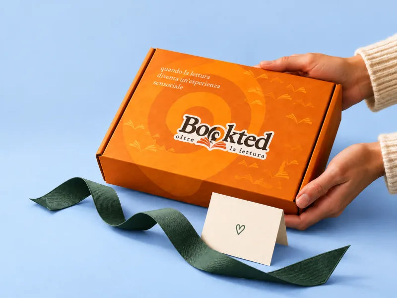 Una mano appoggia una box Bookted arancione, con nastro verde e un biglietto, su un fondo azzurro